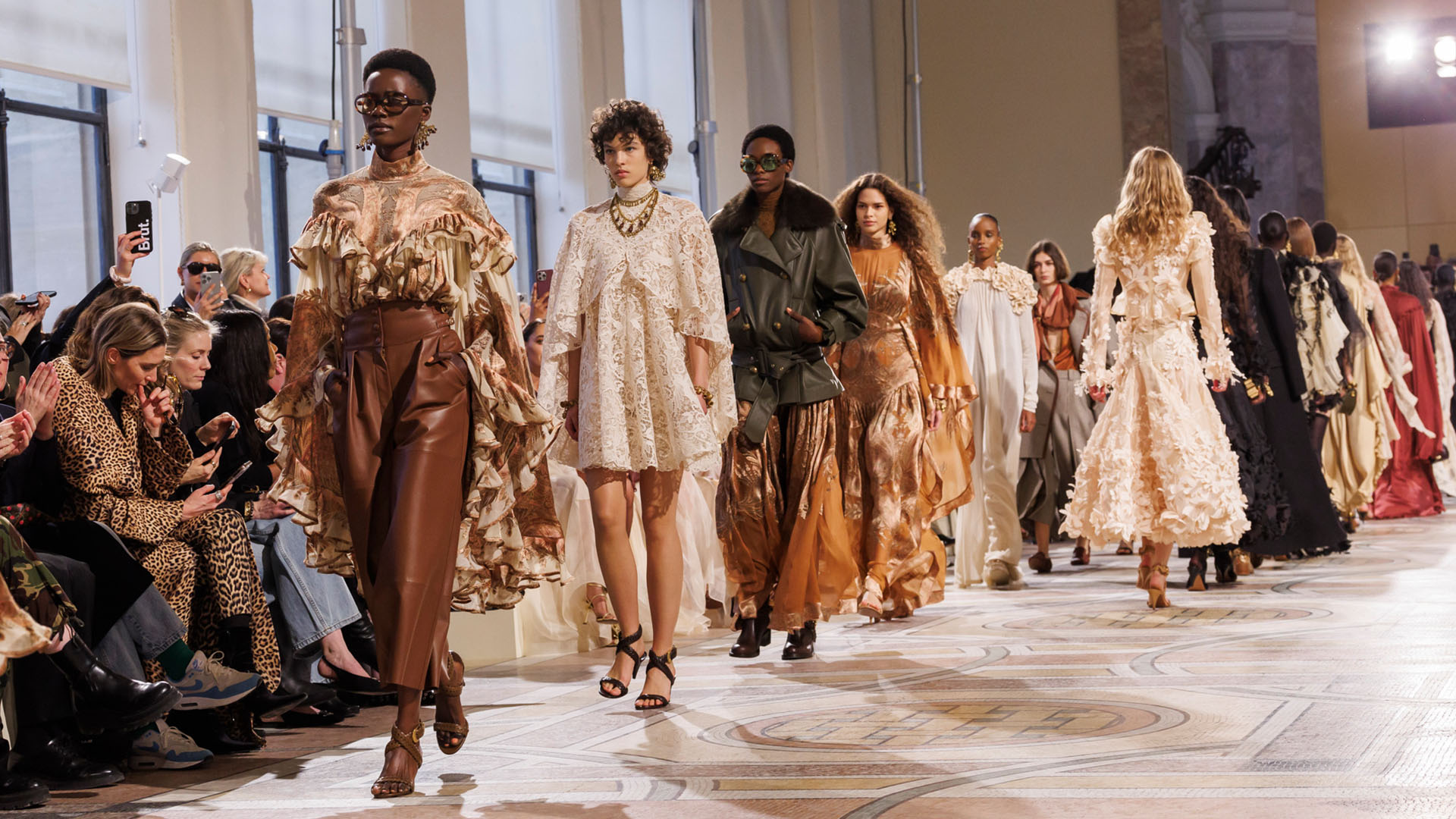 Zimmermann 2025 runway showcasing models in floral lace dresses, bronze silk gowns, leather pants, and statement accessories at a luxury fashion show.